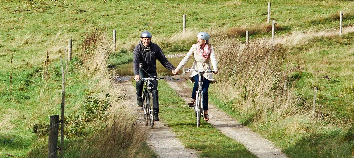 To personer på cykeltur langs Hærvejen ved Gudenåens Udspring cykler hånd i hånd på en forårsdag